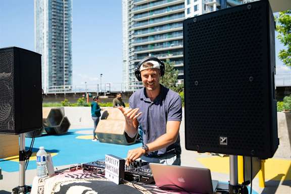 A man in a blue shirt is playing music on a laptop with two large speakers on either side.