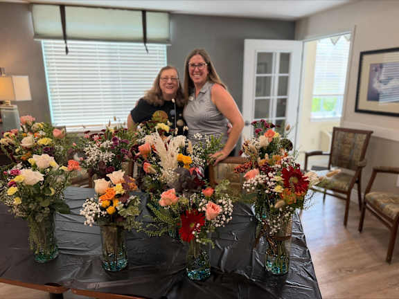 Two women are standing behind a table with a variety of colorful flowers in glass vases.