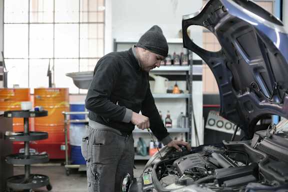 A man in a black beanie and sweater is working on a car engine in a garage.