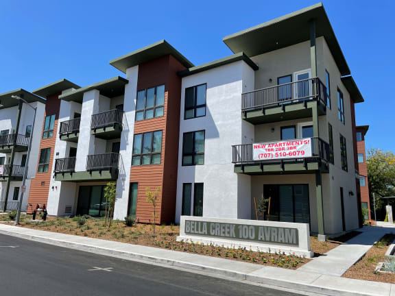 exterior photo of monument sign labeled "100 Avram Ave Bella Creek" in front of exterior of newly constructed apartment building