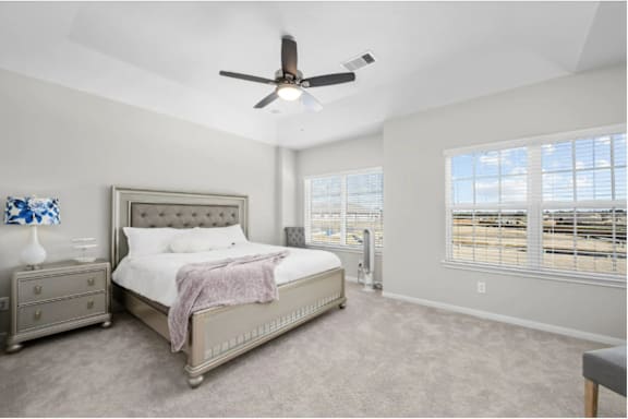A bedroom with a large bed, a ceiling fan, and a window overlooking a parking lot.