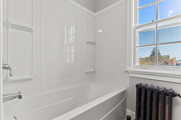 A white bathroom with a tub, window, and radiator.