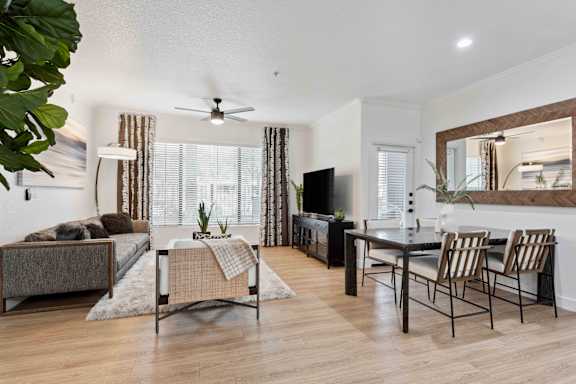 A living room with a brown couch, a grey rug, a wooden floor, a dining table with chairs, a television, and a potted plant.