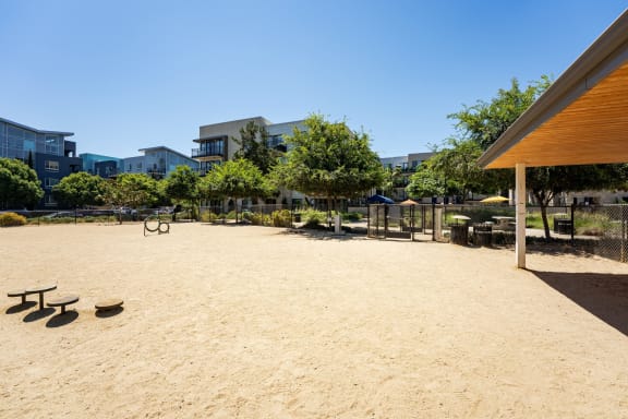 a sandy playground with a pavilion and trees and buildings in the background