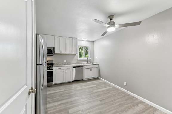 A kitchen with white cabinets and a stainless steel refrigerator, dishwasher, and stove. Window above kitchen sink with blinds. Ceiling fan above dining area.