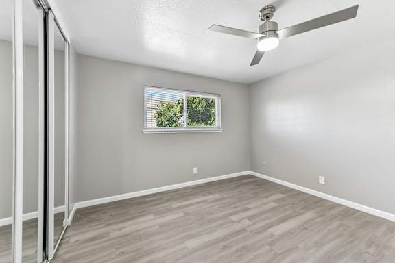 A bedroom with a ceiling fan and a window showing greenery outside. Mirrored closet doors and a open square space.