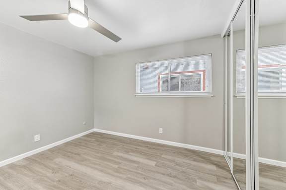 A bedroom with a ceiling fan and a window with blinds. Vinyl plank floors and mirrored closet doors.