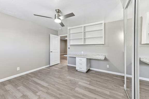 A bedroom with a ceiling fan and a built-in desk with drawers in the middle and shelving above. Vinyl plank flooring and mirrored closet doors.