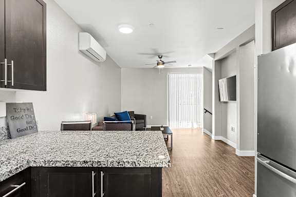 Kitchen view into living room. Bar area with two bar stools, large stainless teel refrigerator, flat screen TV mounted on living room wall, Ceiling fan and mini-split HAVC system in living area. Sliding glass door opposite of kitchen that opens up to balcony with vertical blinds.