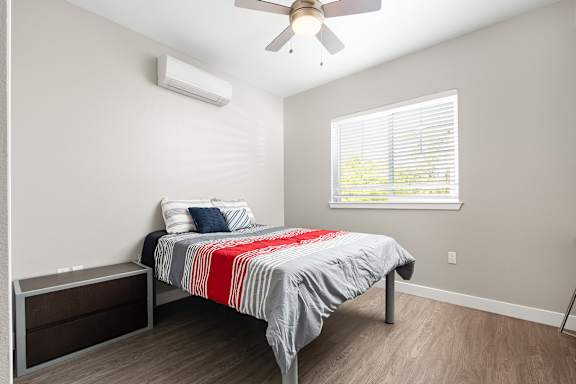 Downstairs bedroom from bedroom door. Full size bed with side table. Ceiling fan and mini-split HVAC system in bedroom. Double window with faux wood blinds. Vinyl plank floors.