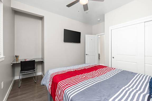 Downstairs bedroom with full size bed, built-in granite desk with black chair, flat screen TV mounted to wall, closet with sliding doors, and ceiling fan and mini-split HVAC system in room.