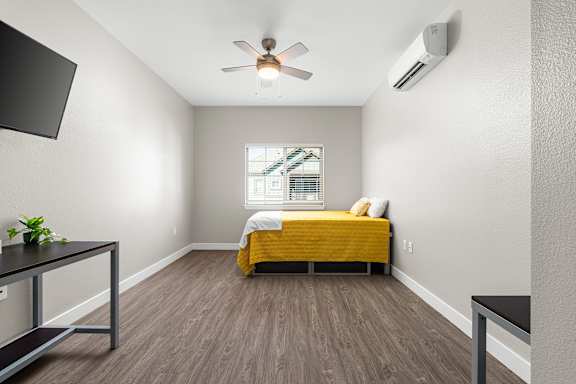 Main bedroom upstairs. Ceiling fan and mini-split HVAC unit in bedroom. Flat screen TV mounted on wall. Full size bed next two a double window. Side table and desk with a modern industrial design of wood and metal. Vinyl plank floors.