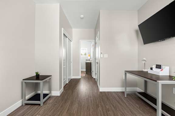 Hall in the main bedroom leading to an attached private bathroom, closet, and bedroom door. Vinyl plank flooring, desk, side table, and flat screen TV mounted to the wall.