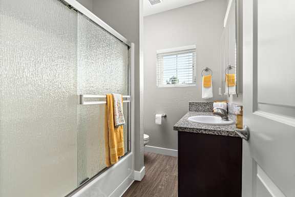 Main bedroom's private bathroom. Frosted glass shower doors with a single basin sink and granite vanity. Window in bathroom with blinds. Vinyl plank floors.