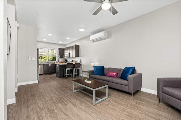Living room with vinyl plank floors, gray couch and oversized gray chair. A wood and metal coffee table in the center of the room. Ceiling fan. Living room is open to the kitchen area and has a mini-split air conditioning unit for the common area.