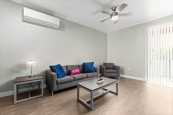 Looking into the living room from the kitchen. Vinyly plank floors, Gray couch, oversized gray chair, coffee table and side table with a modern industrial look. Ceiling fan and mini-split air conditioning unit in living space. Sliding glass door with vertical blinds that opens up to the balcony.