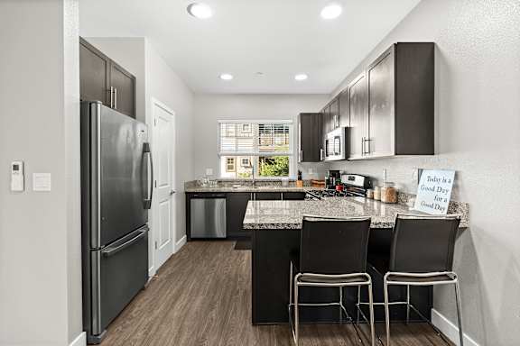 Kitchen view from living room. Large stainless steel refrigerator, dishwasher, and microwave mounted above stove. Espresso colored cabinets and granite counters. Two chairs at the bar. Window above kitchen sink.