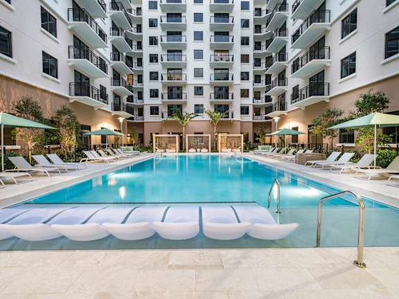A large swimming pool surrounded by lounge chairs and umbrellas in front of a multi-story apartment building.