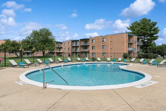 A swimming pool surrounded by lounge chairs and trees in front of a brick apartment building.