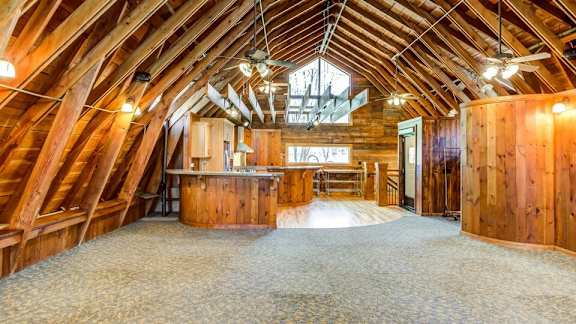 A spacious attic room with wooden walls and ceiling, carpeted floor, and a large window.