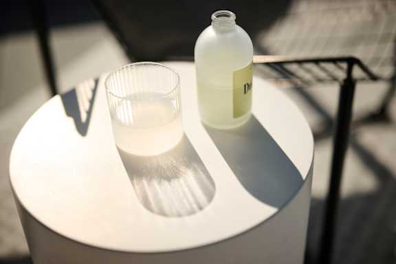 A white bottle with a label that reads "DR" is on a white table next to a glass.