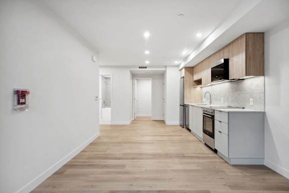 A long, narrow, white hallway with wood floors and a kitchen area on the right.