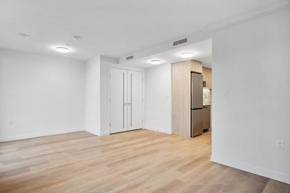 A room with wooden floors and white walls, with a kitchenette in the corner.