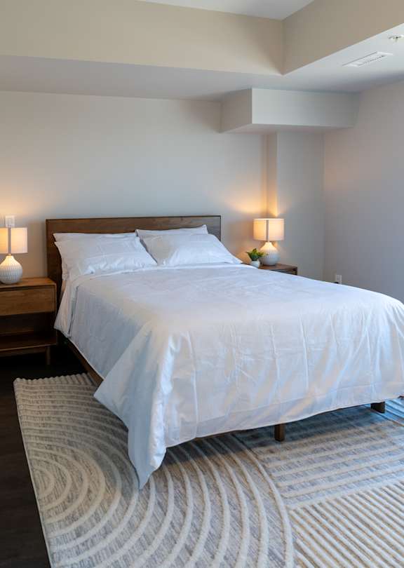 A bed with white bedding sits in a room with a striped rug.
