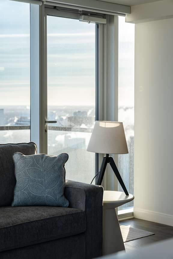 A grey couch with a blue pillow is in front of a window with a city view.