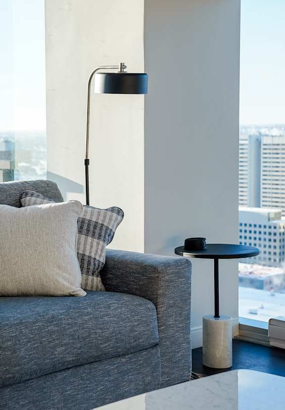 A grey couch with a white pillow and a black table with a lamp on it.