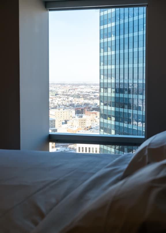 A bed with white sheets sits in front of a window with a view of a cityscape.