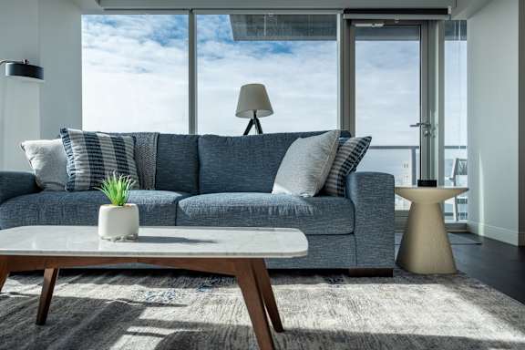 A grey couch with a white vase on a wooden table in front of a window.