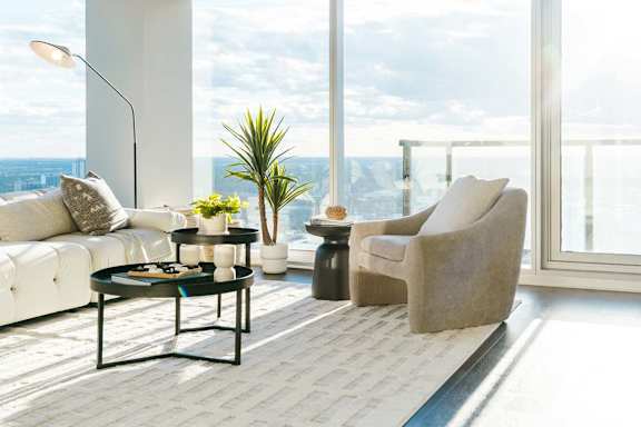 A living room with a white sofa, a beige armchair, a black coffee table, and a large window overlooking the city.