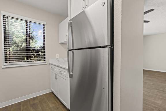 A stainless steel refrigerator in a kitchen with wooden flooring and a window with blinds.