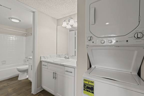 A white washing machine is in a laundry room with a sink and mirror.
