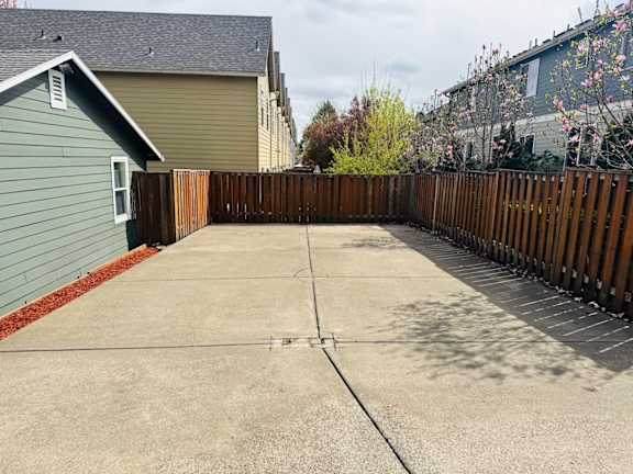 A concrete patio with a wooden fence and a green house in the background.