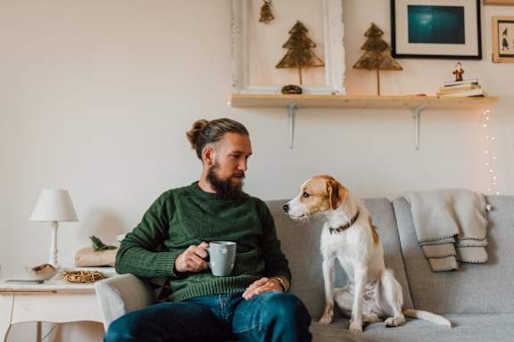 A man with a beard is sitting on a couch with a cup in his hand and a dog standing next to him.