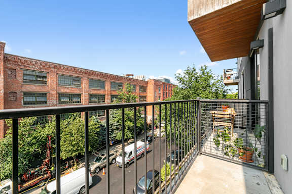 A balcony with a black railing overlooks a parking lot and a brick building.