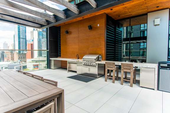A wooden table is in the middle of a patio with a grill and chairs.