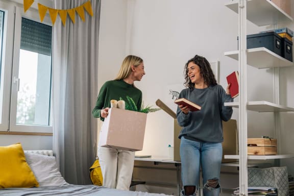 two women standing in a bedroom holding a box and a book