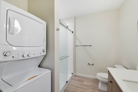 A white washing machine is in a small laundry room with a toilet and sink.