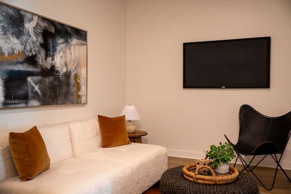 A living room with a white couch, a black chair, a small table with a plant and a basket on it, and a large painting on the wall.