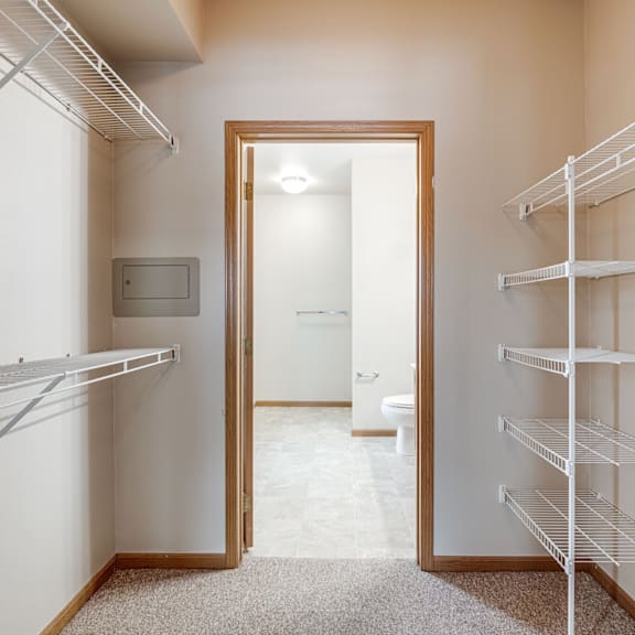 A white closet with shelves and an open door leading to a bathroom.