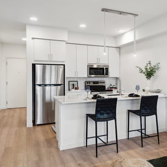 A modern kitchen with a white island, two black chairs, and a stainless steel refrigerator.