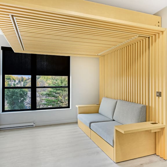 A wooden sofa with grey cushions sits under a wooden beam in a room with a window.