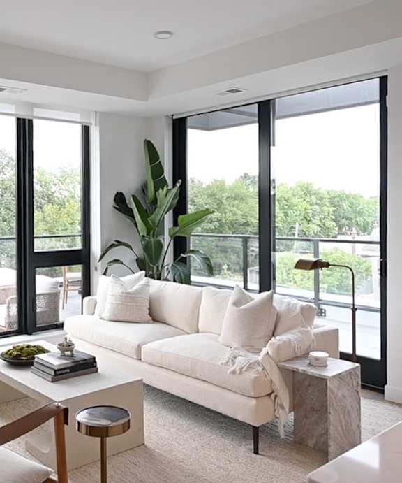 A white couch with pillows and a plant on a table in front of a window.