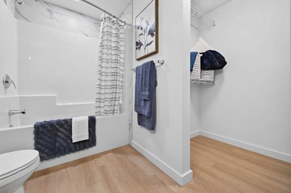A bathroom with a white toilet, a white sink, a white bathtub, a white shower curtain, a white towel, a blue towel, a blue bag, a white shelf, a white wall,