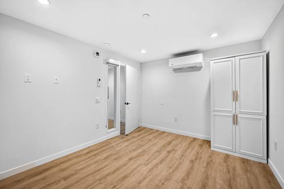 A room with a wooden floor and white walls, with a door on the right and an air conditioner on the wall.