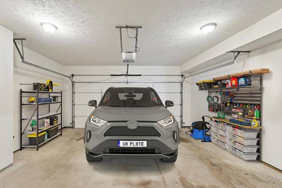 A grey car is parked in a garage with a white wall and a shelf with tools.