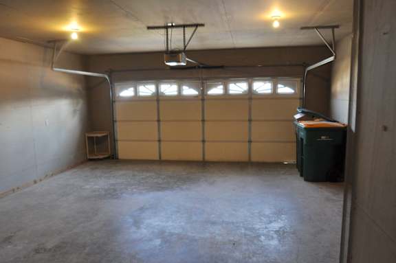 A large empty garage with a closed door and a green trash can.